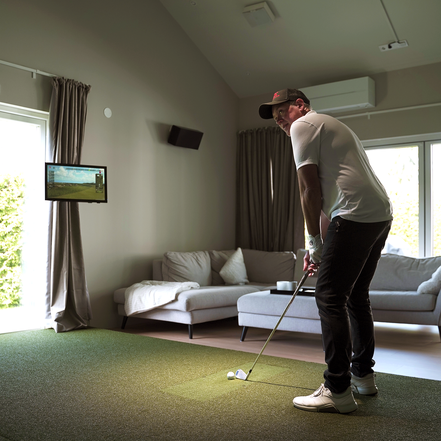 Man spielt Golf mit einem Simulator im Wohnzimmer, steht auf Kunstrasen mit einem Schläger, Sofa und TV-Bildschirm im Hintergrund.