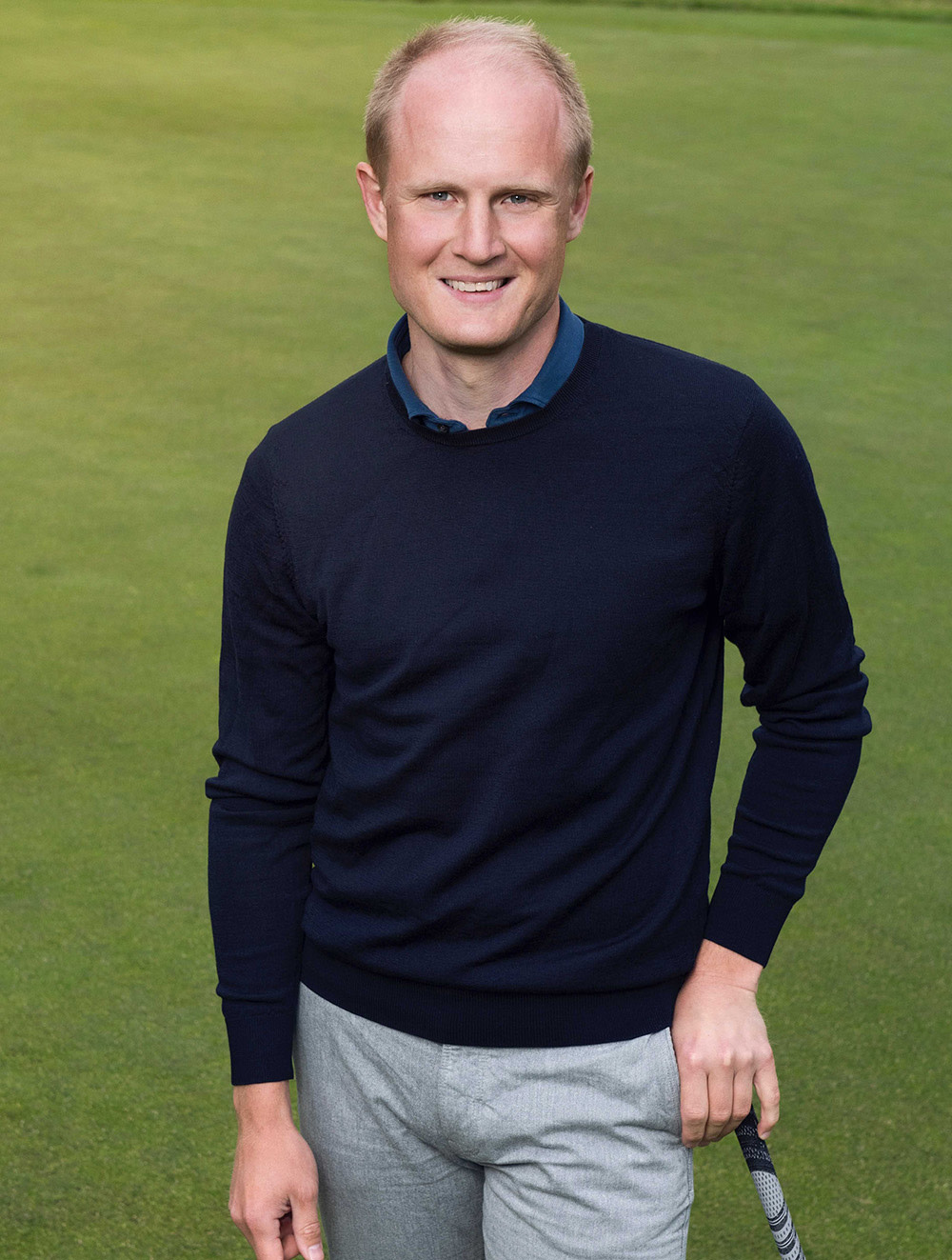 Man i mörkblå tröja och grå byxor står på en golfbana och håller i en golfklubba, leende mot kameran.