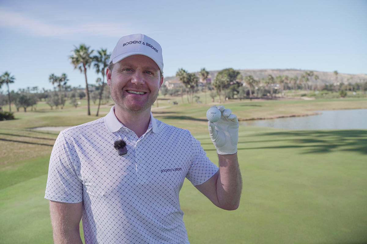 Leende man i golfkläder håller upp en golfboll på en golfbana med palmer och vatten i bakgrunden.
