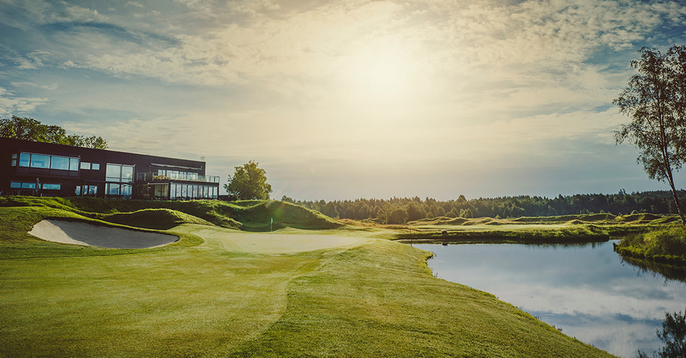Golfbana med green, sandbunker och vattenhinder i solnedgång, klubbhus till vänster och skog i bakgrunden.