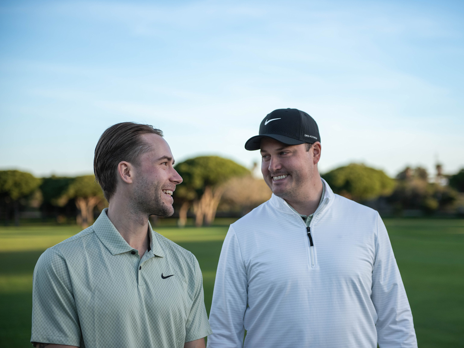 Kaksi miestä seisoo ulkona golfkentällä, hymyilevät ja katsovat toisiaan, molemmilla on yllään Nike-vaatteet.