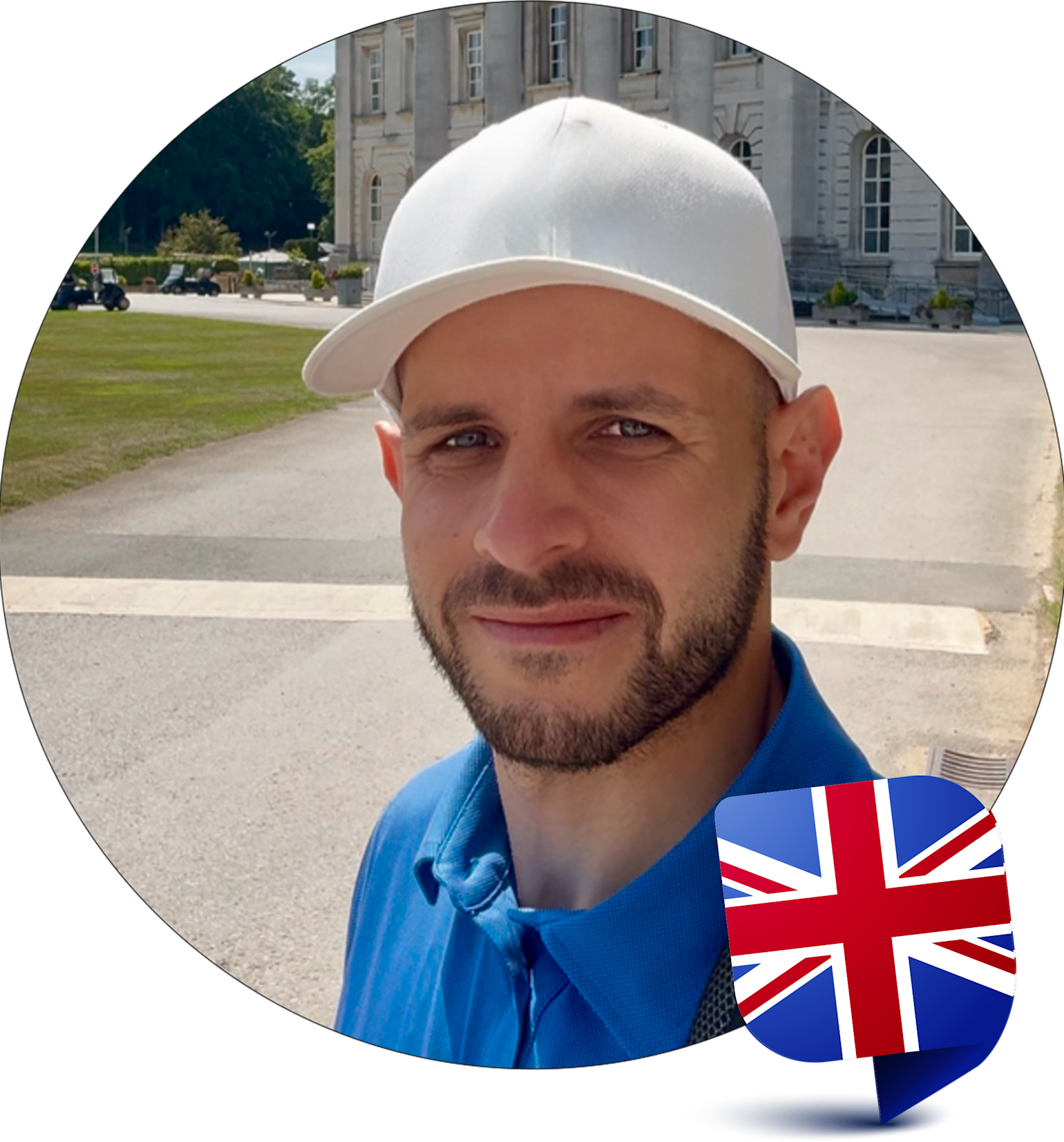 Mann mit Bart und weißer Mütze lächelt im Freien vor einem Gebäude, mit britischer Flagge in der unteren rechten Ecke.