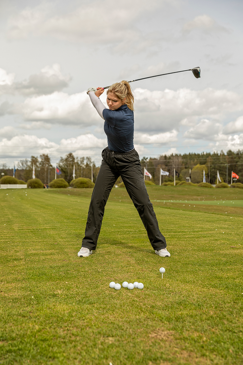 Kvinna svingar en golfklubba på en driving range, med flera golfbollar på gräset framför sig och flaggor i bakgrunden.