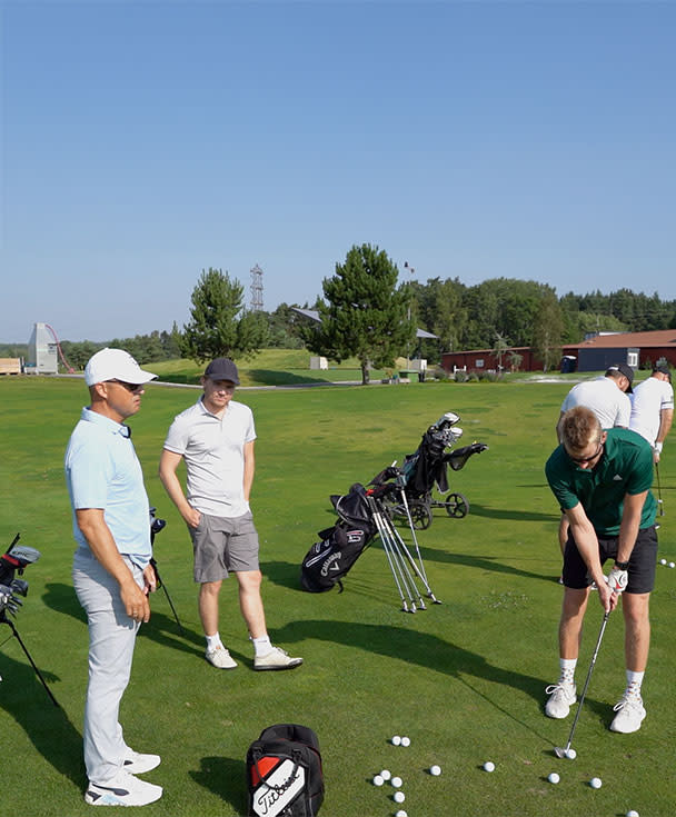 Tre män står på en golfbana, en av dem svingar en golfklubba medan de andra tittar på, golfbagar och bollar syns på gräset.