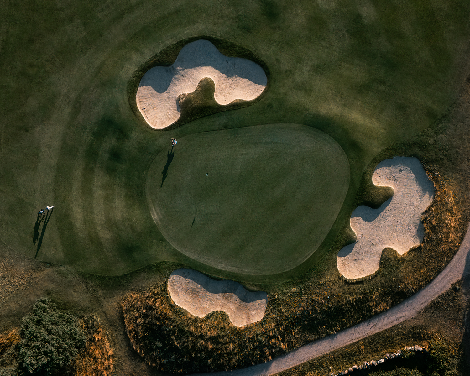 Två golfare går på en green omgiven av tre sandbunkrar, fotograferat ovanifrån i kvällsljus.