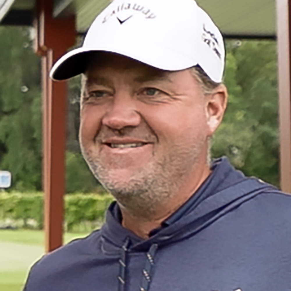 Man med vit Callaway-keps och blå huvtröja ler utomhus på en golfbana med gröna träd i bakgrunden.