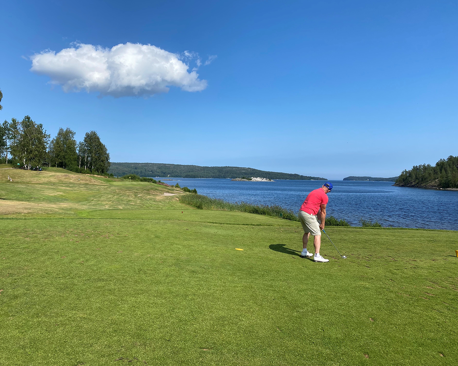 En man i röd tröja och ljusa shorts slår en golfboll på en grön golfbana vid en sjö med blå himmel och en vit moln.