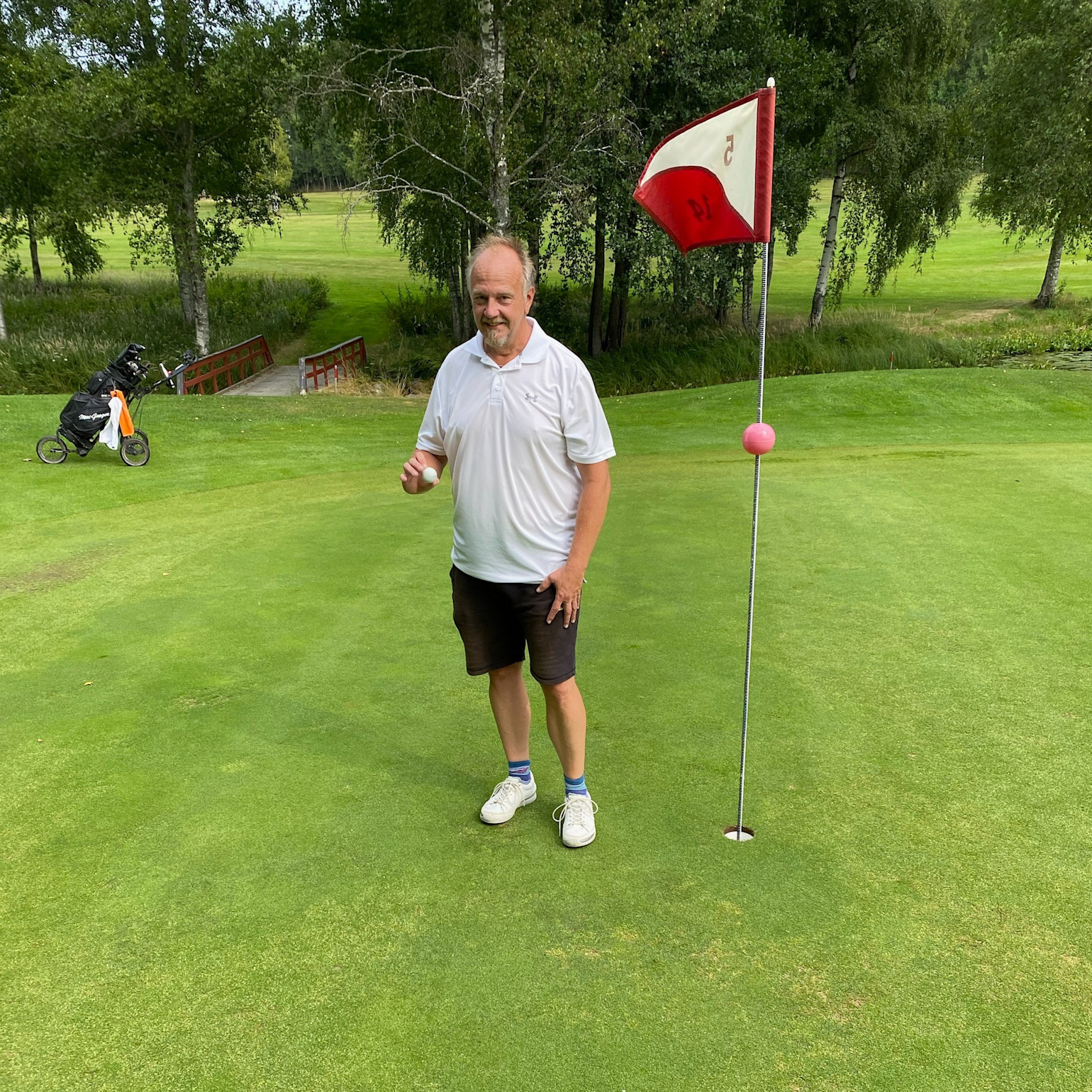 Mand iført hvid poloshirt og sorte shorts står ved siden af et golfhul med flag på en golfbane og holder en golfbold.