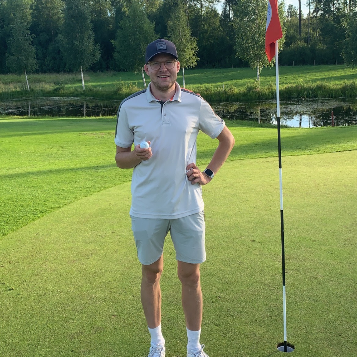 En mand i golfbeklædning står ved siden af et hul med flag på en golfbane, holder en golfbold og smiler til kameraet.