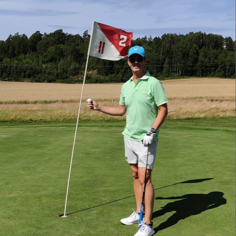 Mand iført grøn poloshirt og blå kasket holder en golfbold og står ved flag nummer 2 på en golfbane med skov i baggrunden.