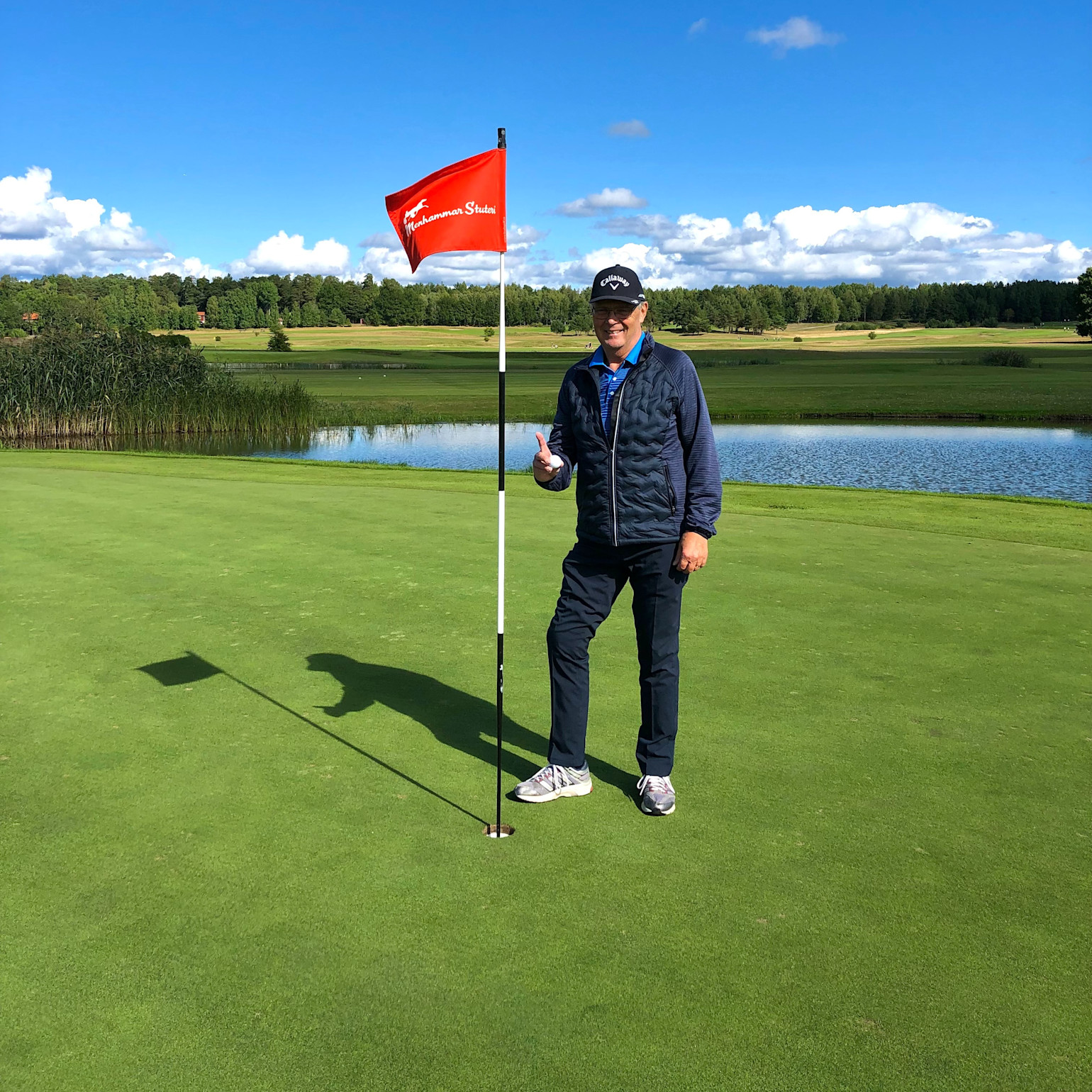Man står ved siden af et golf-flag på greenen, giver tommelfingeren op, med sø og træer i baggrunden under blå himmel.