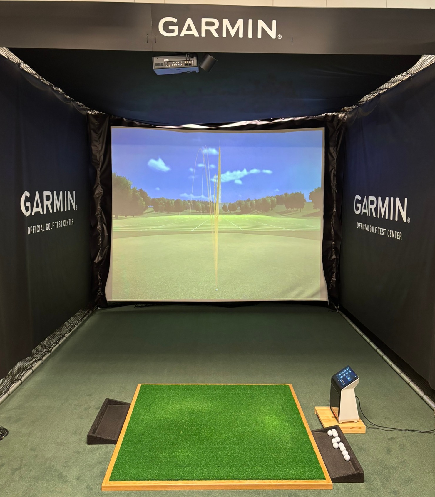 Garmin golf-simulator med konstgräsmatta, golfbollar och digital skärm som visar golfbanan och bollens bana.
