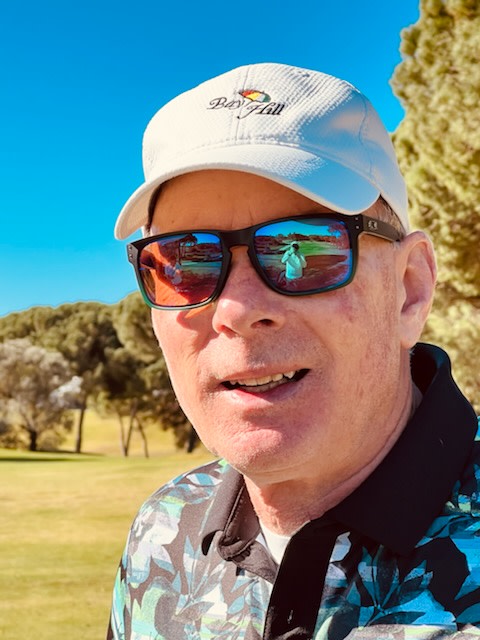 Mand med solbriller og hvid kasket med teksten "Bay Hill" står udendørs på en golfbane med træer og blå himmel i baggrunden.