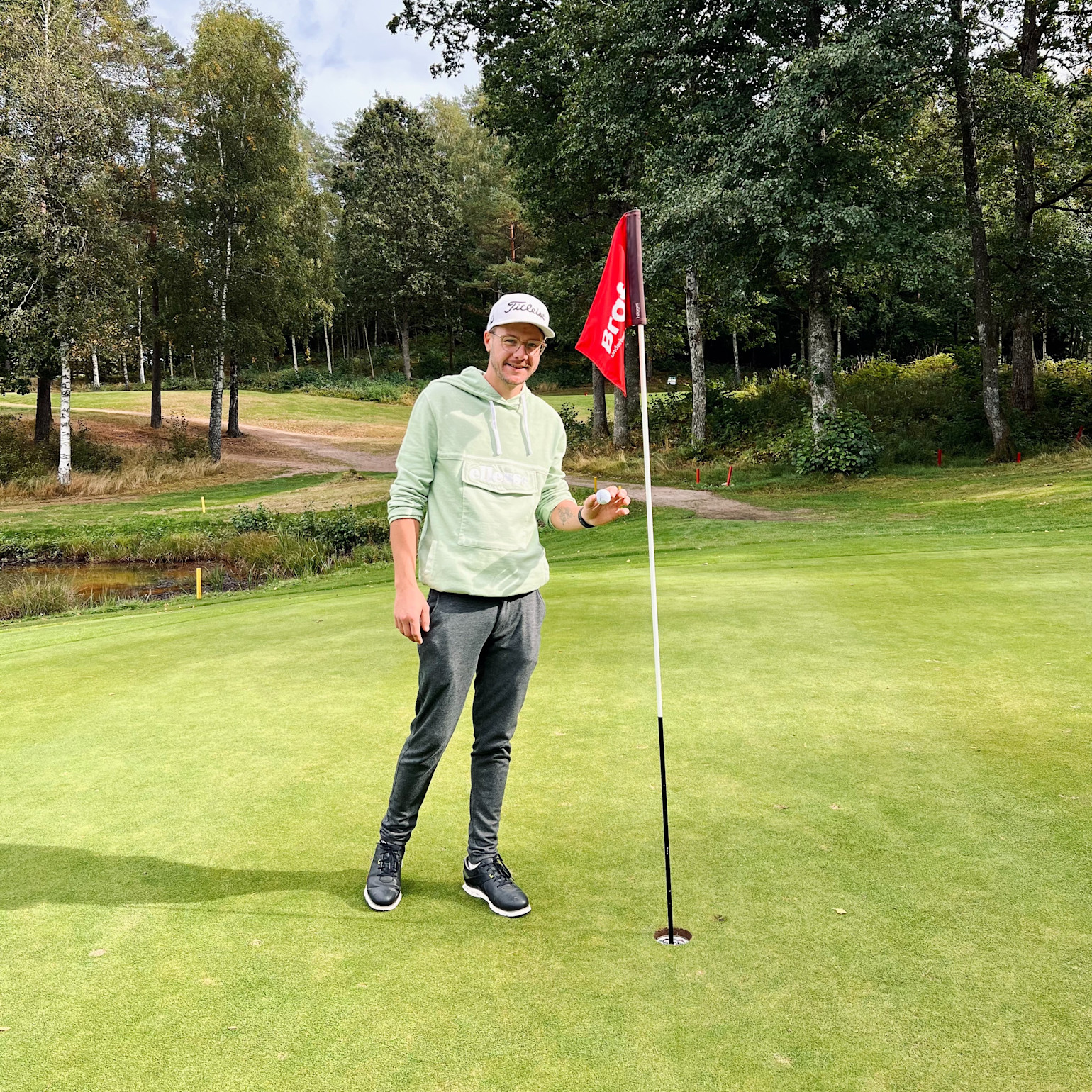 Man i ljusgrön hoodie och keps håller en golfboll bredvid ett hål med röd flagga på en golfbana omgiven av träd.