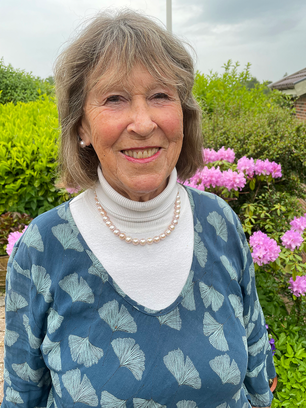 Ældre kvinde med gråt hår og perlekæde smiler udendørs, omgivet af grønne buske og lyserøde blomster.