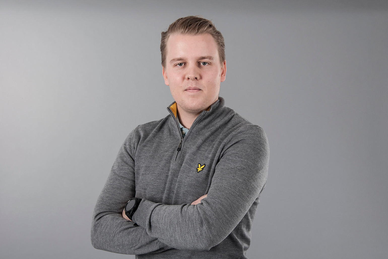 Man med armarna i kors bär grå tröja med gul logotyp och svart klocka, står mot ljusgrå bakgrund.
