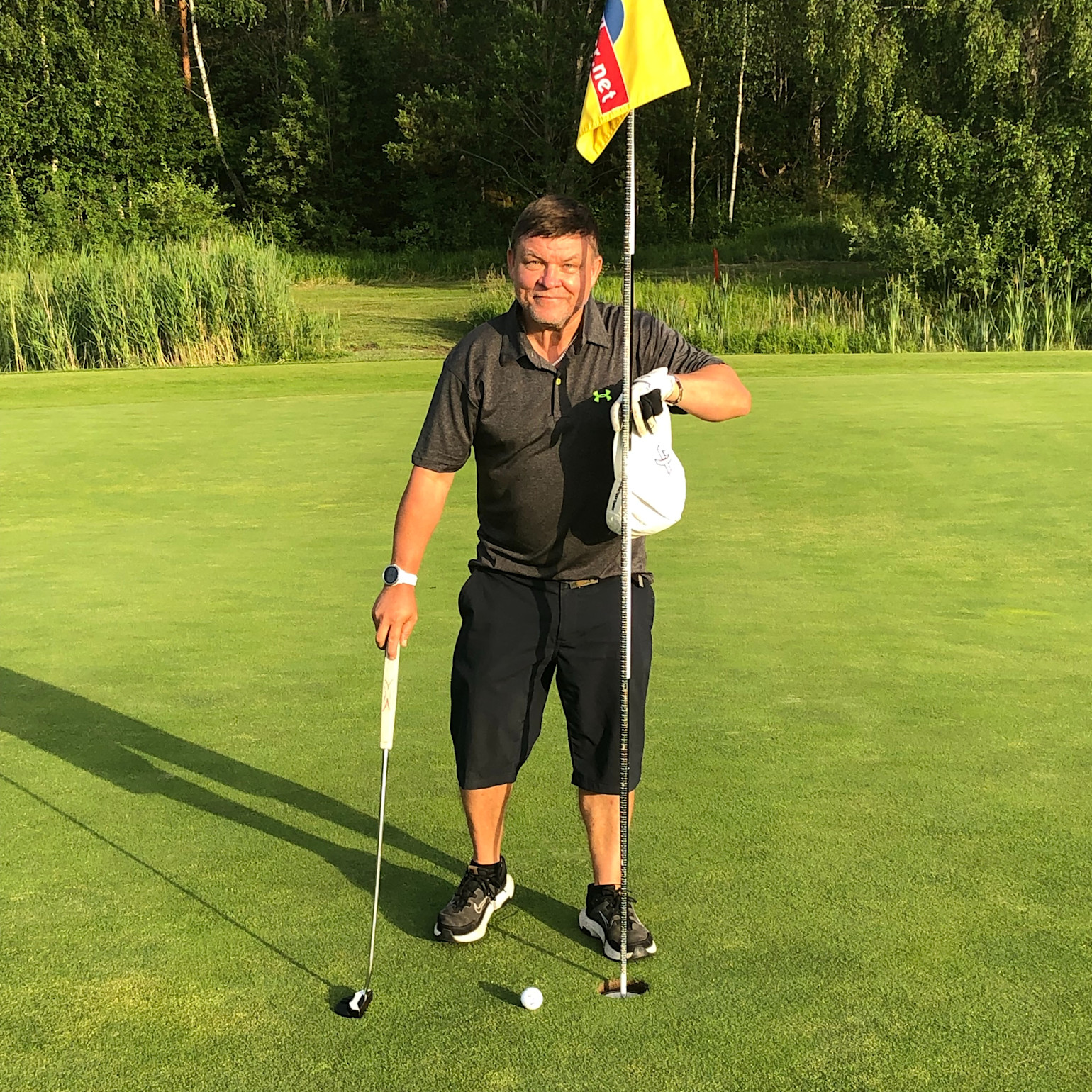Man spiller golf, holder i flagstangen og en golfkølle på greenen, med en golfbold tæt på hullet og skov i baggrunden.