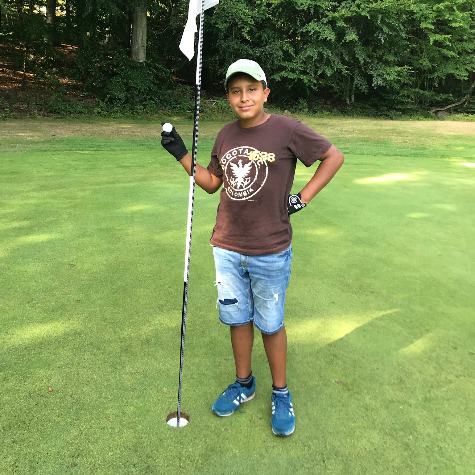 Dreng med kasket og golfhandske holder en golfbold ved siden af flagstangen på greenen, omgivet af grønne omgivelser.