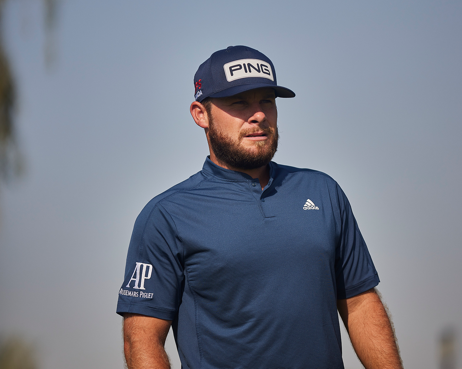 Man med skägg bär blå PING-keps och blå Adidas-tröja med Audemars Piguet-logotyp utomhus under klar himmel.