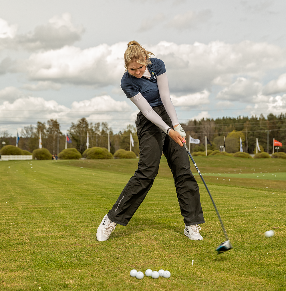 Eine Frau schwingt einen Golfschläger und schlägt einen Golfball auf einer grünen Driving Range, auf der mehrere Bälle auf dem Boden liegen.