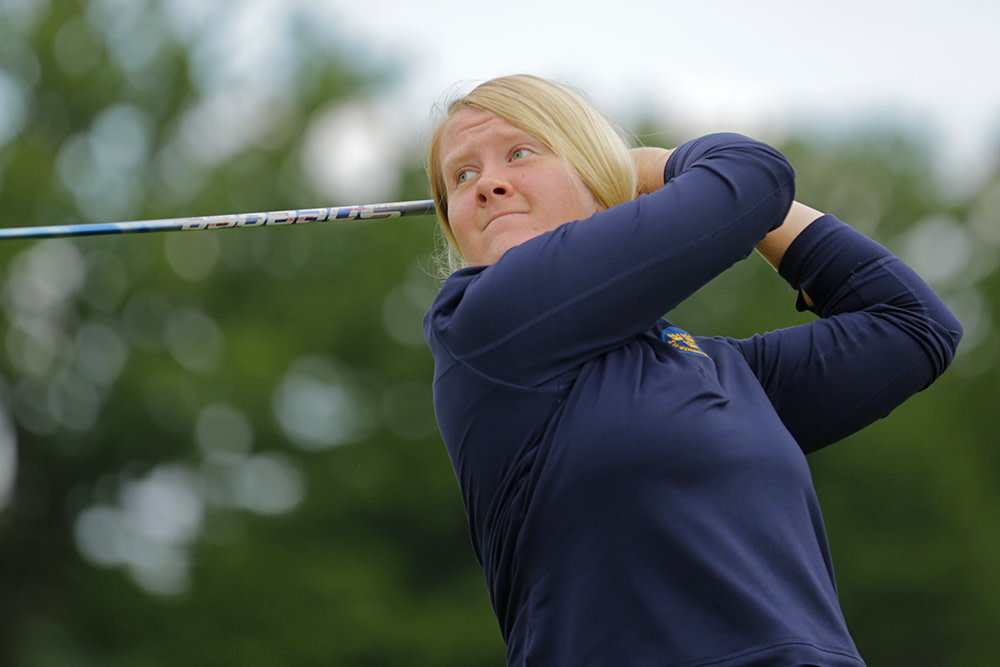 Kvinna med blont hår svingar en golfklubba utomhus, klädd i mörkblå tröja med suddig grön bakgrund.