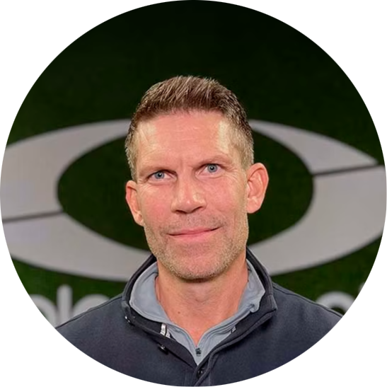 Man med kort brunt hår ler mot kameran, står framför en vägg med en stor oval logotyp i bakgrunden.