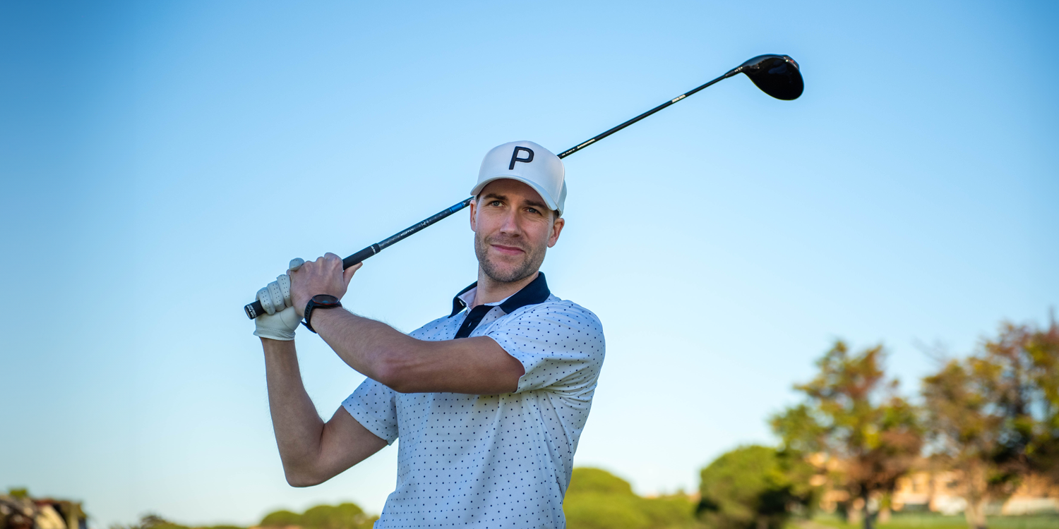 Ein Mann mit weißer Kappe mit dem Buchstaben P hält einen Golfschläger über den Schultern auf einem Golfplatz unter klarem, blauem Himmel.