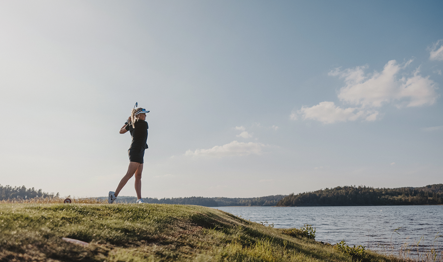 Kvinne svinger en golfkølle på en grønn golfbane ved vannet under en klar blå himmel.