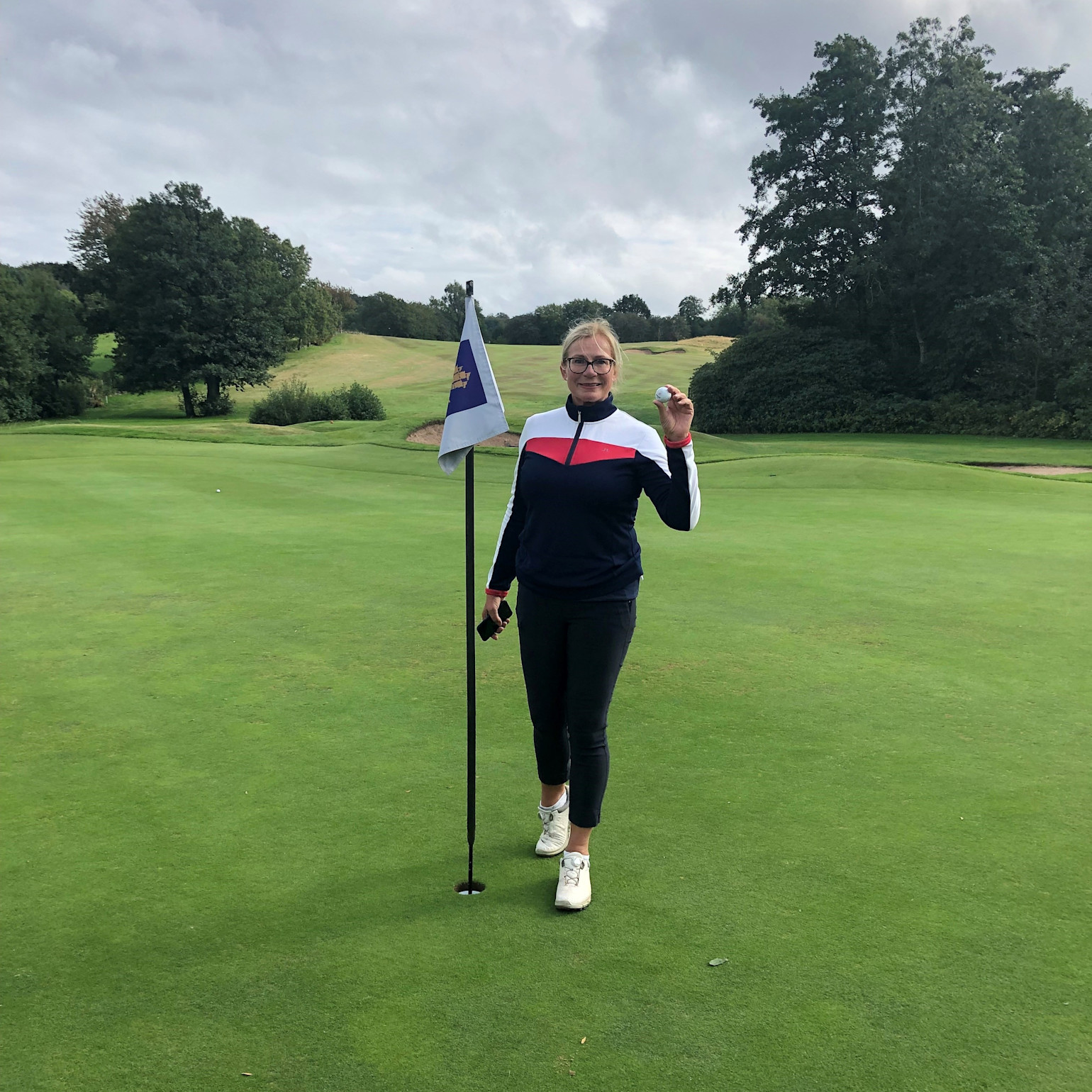 Kvinde står ved siden af et golf-flag på en green og holder en golfbold op, omgivet af en frodig golfbane og træer.