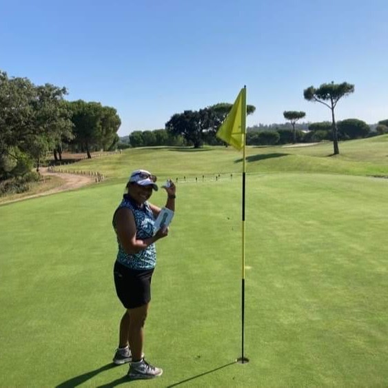 Kvinde står ved siden af et golf-flag på greenen, holder en golfbold op og smiler på en solrig golfbane.