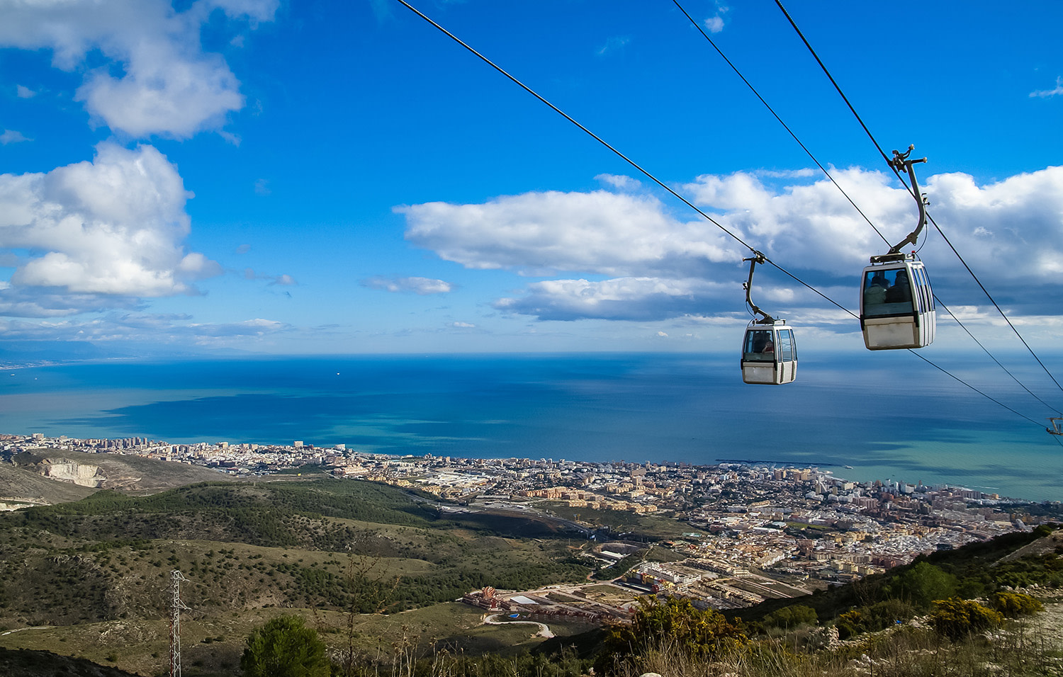 Två linbanegondoler svävar över en stad vid kusten med hav och blå himmel i bakgrunden.