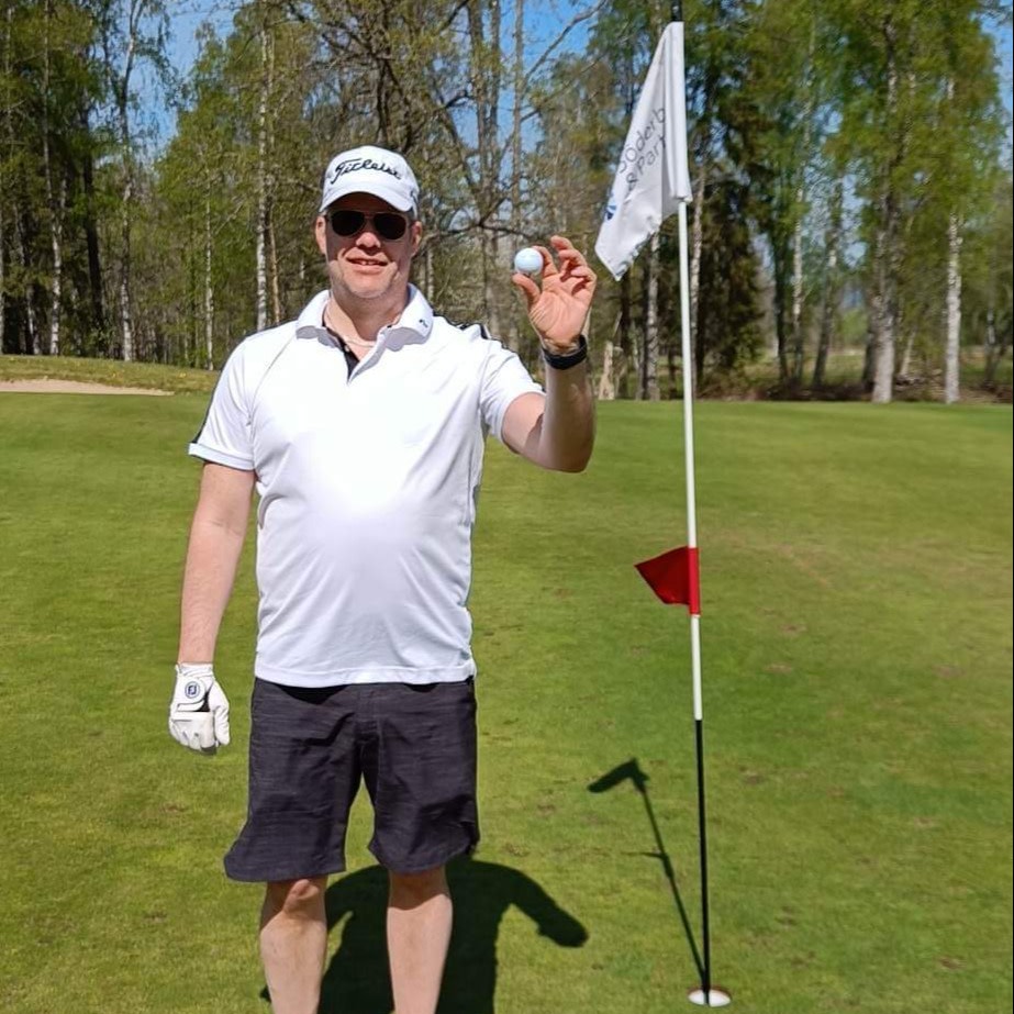 Mand i hvid poloshirt og kasket holder en golfbold op ved siden af en flagstang på en golfbane med grønne omgivelser i baggrunden.