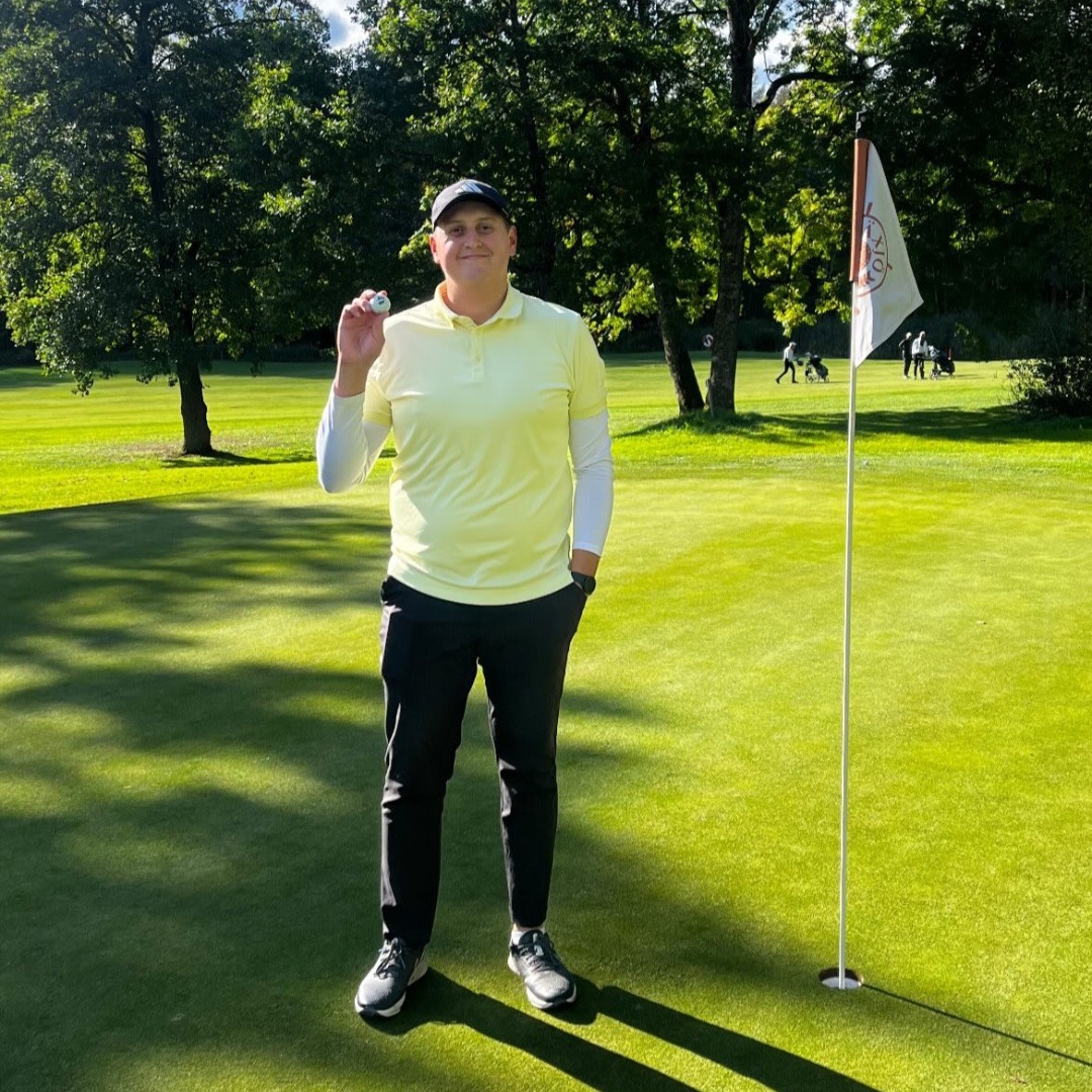 Mand i gul trøje står på en golfgreen, holder en golfbold op ved siden af flaget, med træer og golfspillere i baggrunden.