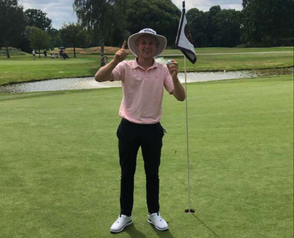 Mand i lyserød polo og solhat holder en golfbold op og peger opad ved siden af et flag på en golfgreen.