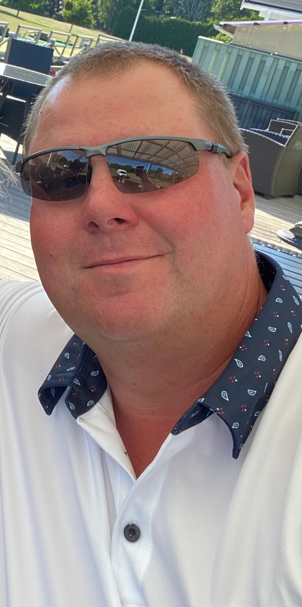 Mand med kort hår og solbriller smiler og bærer en hvid poloshirt med mønstret krave udendørs på en terrasse.