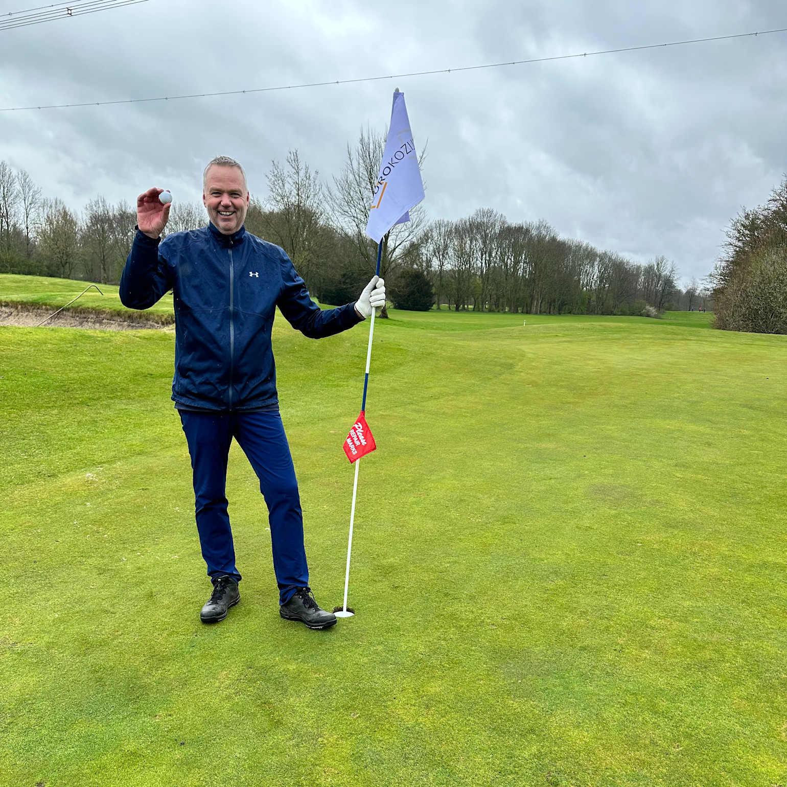 Mand i blå jakke holder en golfbold og en flagstang på en golfbane med grønt græs og træer i baggrunden under en overskyet himmel.