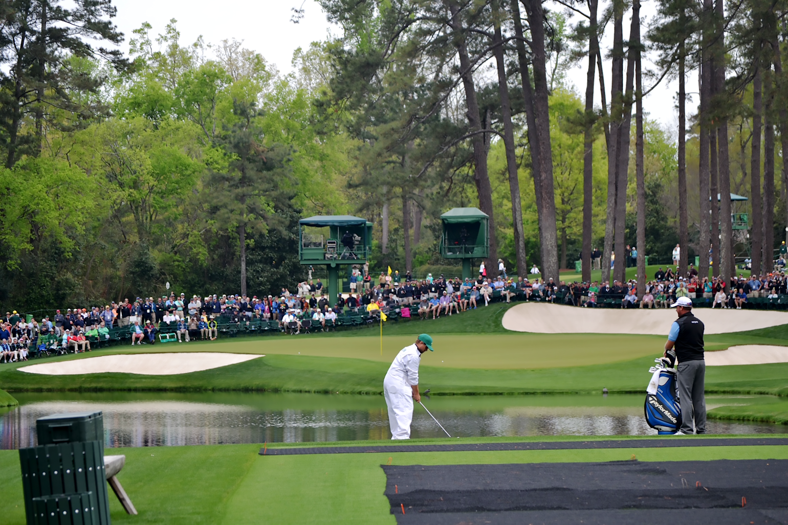 Golfspelare slår ett slag över en damm mot greenen, med publik och höga träd i bakgrunden under en golftävling.