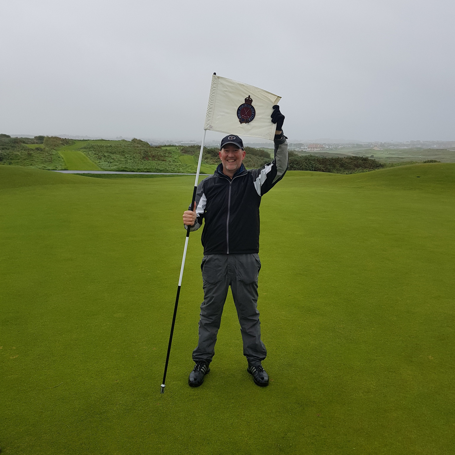 En mand iført golfbeklædning holder en flagstang og smiler på en golfbane med grønt græs og tåget baggrund.