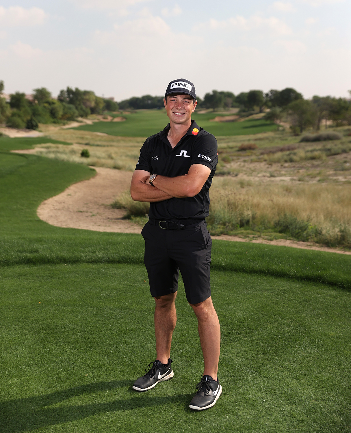 Man i svart golfkläder och keps står med armarna i kors på en golfbana med grönt gräs och träd i bakgrunden.