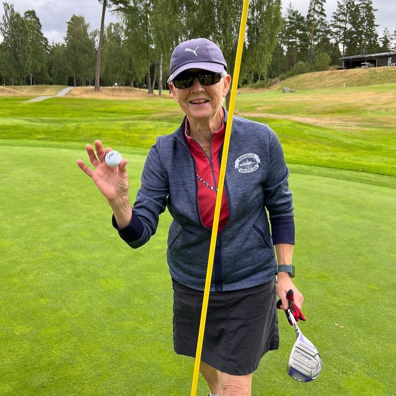 Smilende kvinde med solbriller og kasket holder en golfbold op ved siden af flagstangen på en green.