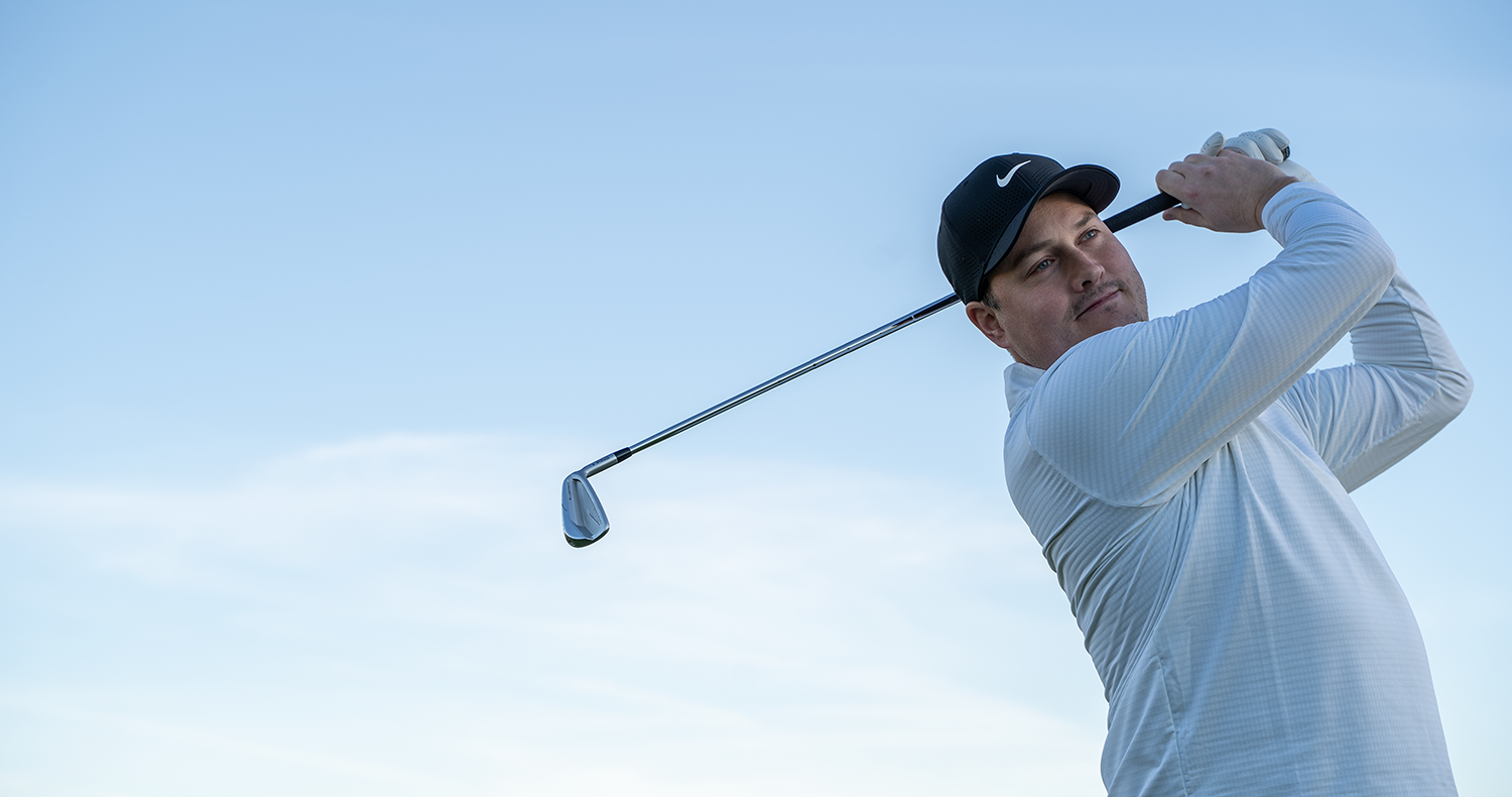 Man i vit tröja och svart keps svingar en golfklubba utomhus mot en klarblå himmel.