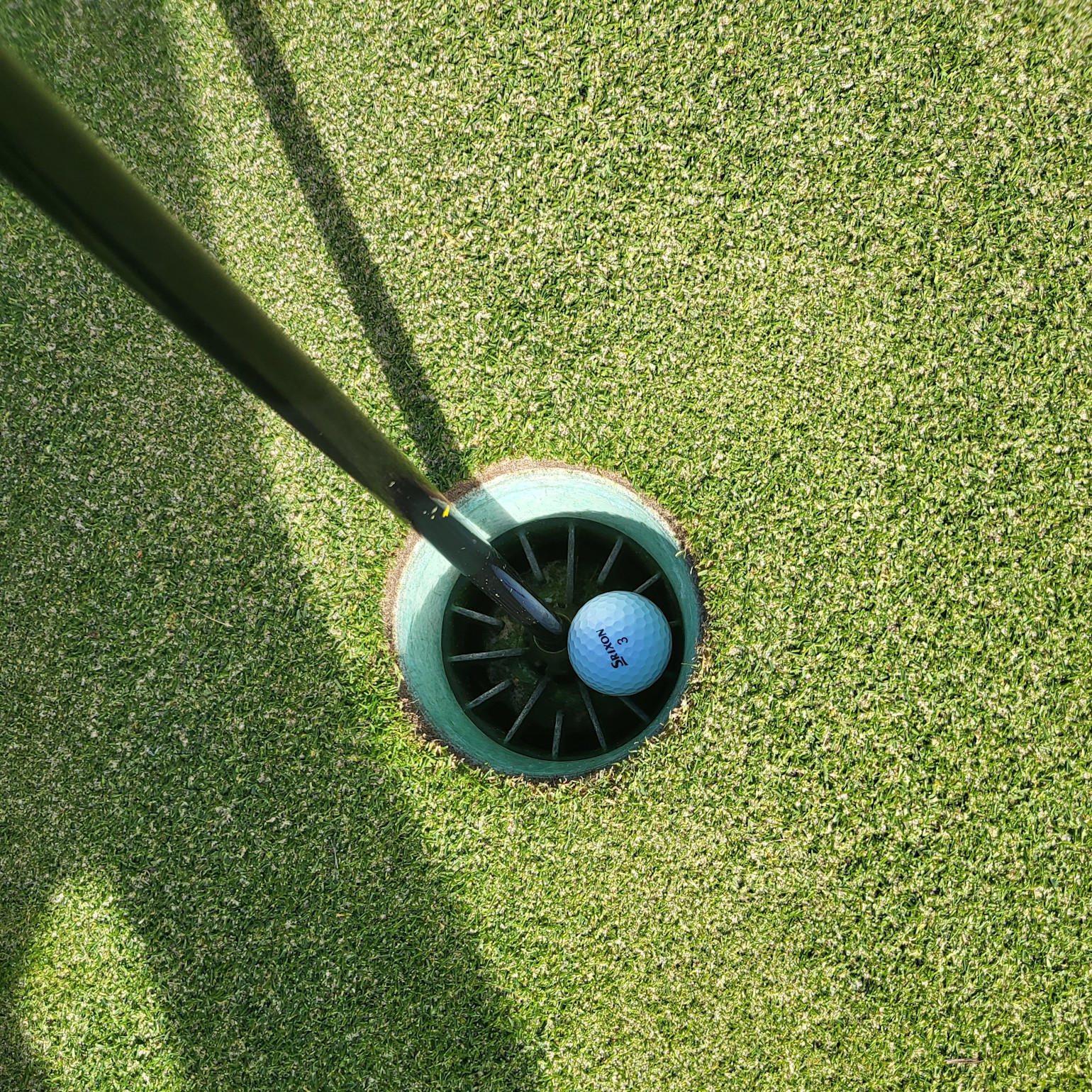 En golfbold ligger i hullet ved siden af flagstangen på en velplejet green under solskinsvejr.