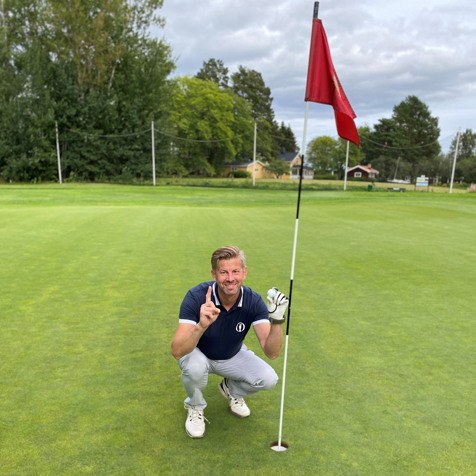 Mand iført golfbeklædning huker sig ned ved siden af et golfhul med flag, mens han holder en finger og en golfbold op på en grøn golfbane.