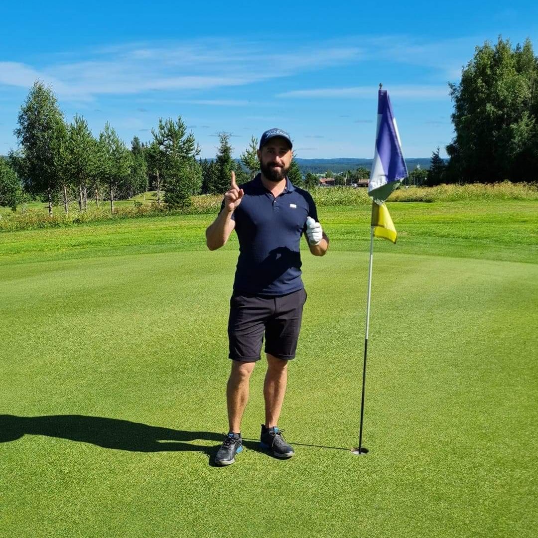 En mand iført golfbeklædning står ved siden af et flag på en golfgreen, løfter en finger og smiler, med træer og blå himmel i baggrunden.