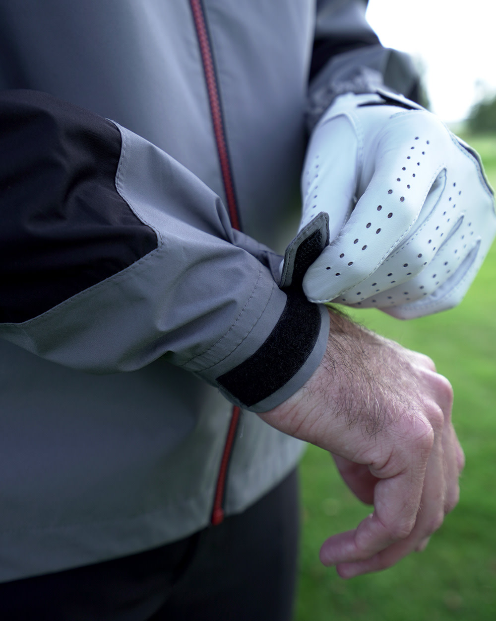 Person justerar kardborrebandet på ärmen av en grå jacka, bär vit golfhandske, utomhus på gräs.