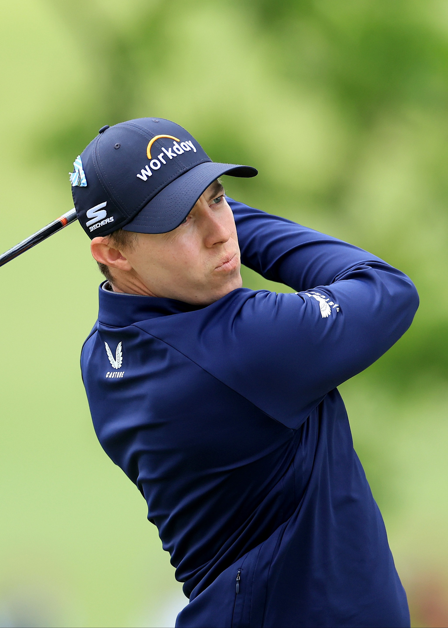 Golfspieler schwingt einen Schläger auf einem Golfplatz, trägt eine blaue Kappe mit der Aufschrift „workday“ und eine blaue Jacke.