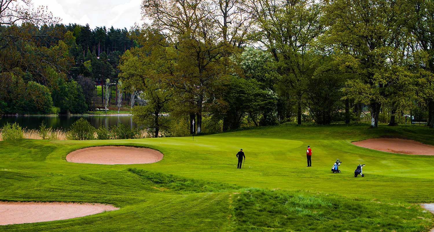 Två golfare står på en grön golfbana omgivna av träd, med golfbagar och sandbunkrar i närheten.