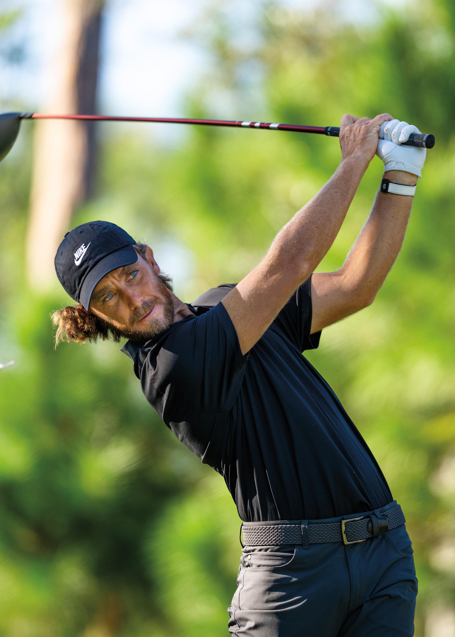 Mann in schwarzem Poloshirt und Mütze schwingt einen Golfschläger auf einem Golfplatz mit grüner, blühender Kulisse.