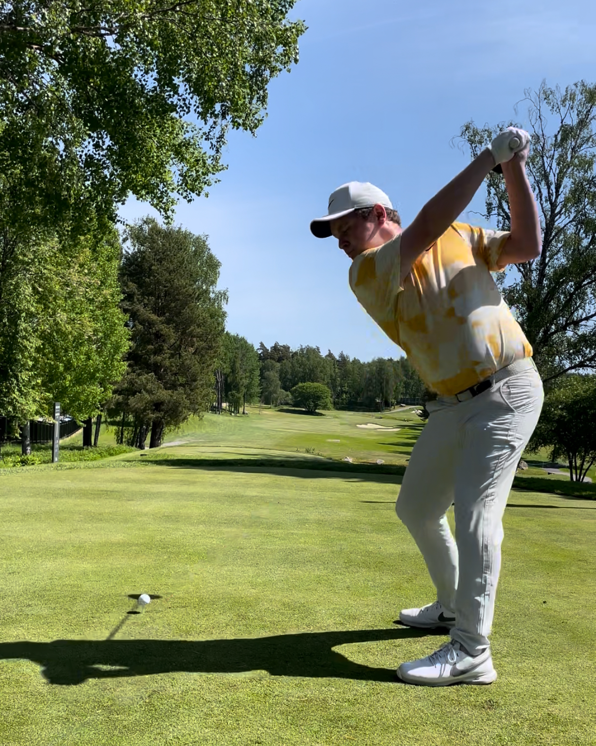 Ein Golfspieler in einem gelb-weißen Hemd und einer weißen Kappe schwingt einen Schläger auf einem grünen Golfplatz, der von Bäumen umgeben ist, unter klarem, blauem Himmel.