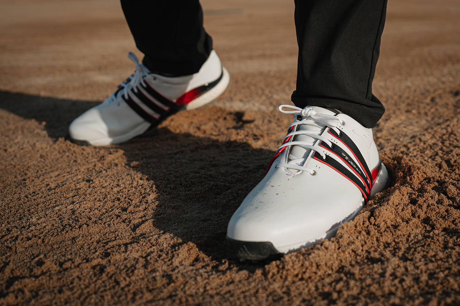 Vita sportskor med svarta och röda detaljer används av en person som står på sandig mark, troligen på en golfbana.